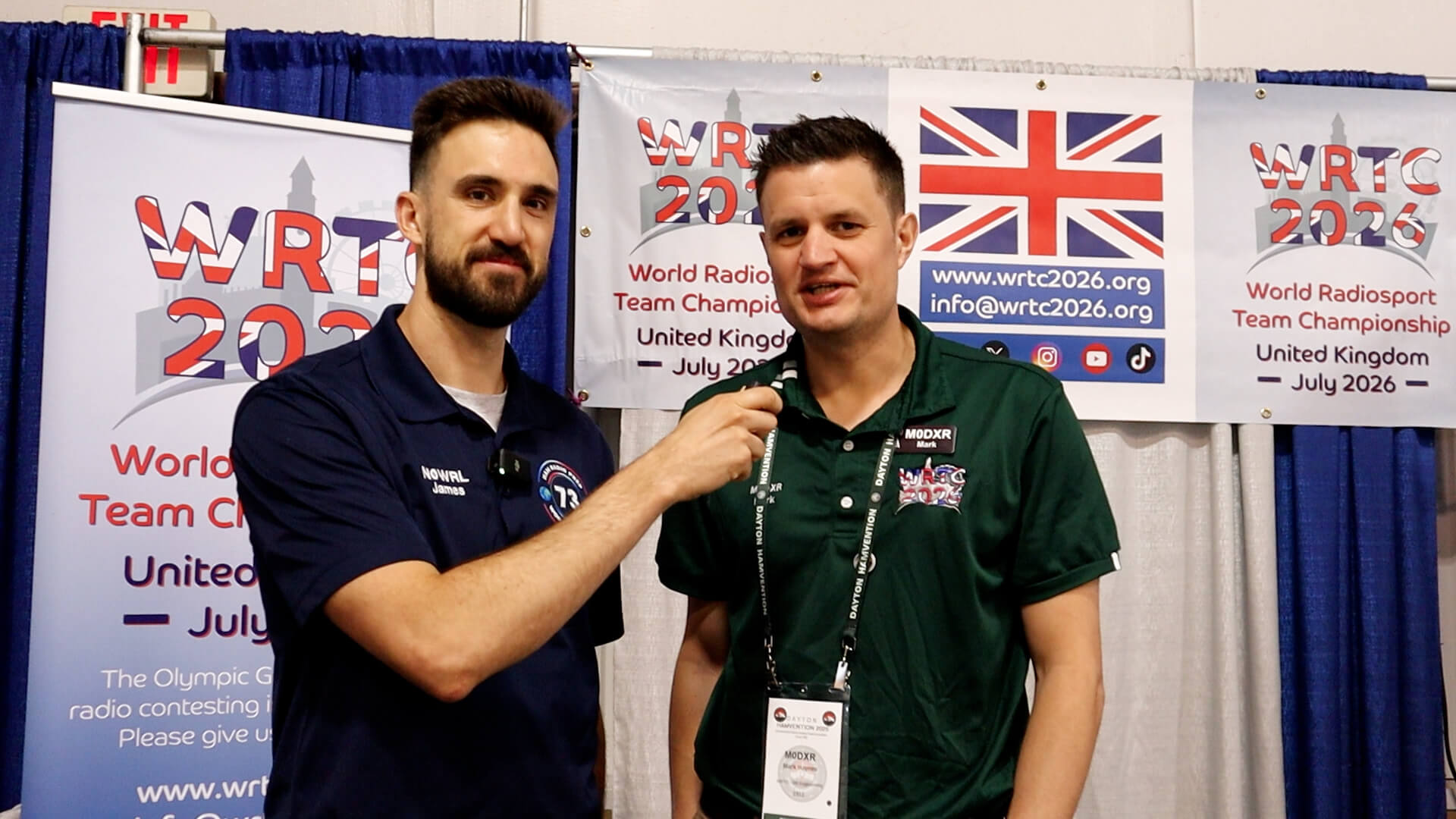 Two people at a WRTC 2026 booth; the person on the left holds a microphone toward the person on the right, with UK and event banners in the background.